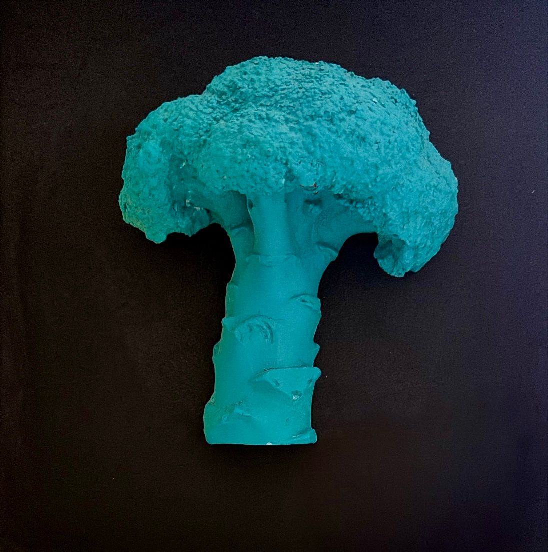 Un broccolo modellato in pasta azzurra, su un fondo nero.