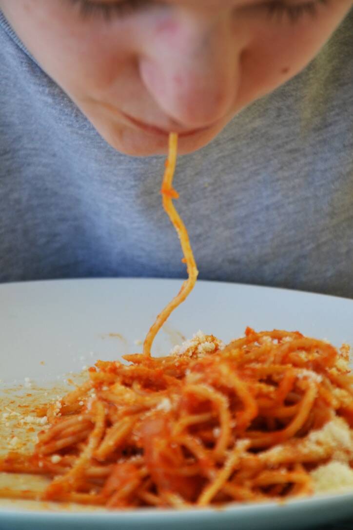 Una ragazza raccoglie con la bocca un lungo filo di spaghetti al pomodoro da un piatto bianco.